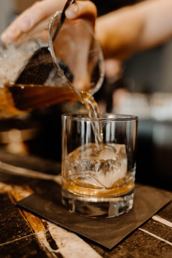 A hand holding a glass of whiskey with an orange slice garnish a bottle of Dread River whiskey is blurred in the background.