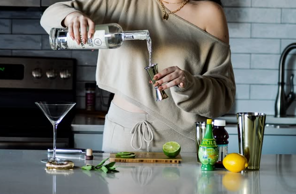 A woman in a beige lounge set pours clear liquor into a jigger at a kitchen counter, surrounded by a martini glass, shaker, lemons, and limes.