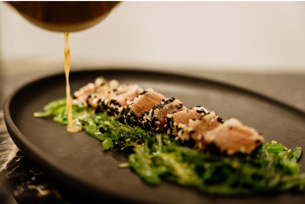 A close-up photograph of a gourmet dish featuring sesame-crusted seared tuna slices arranged in a row on a bed of fresh green seaweed salad.
