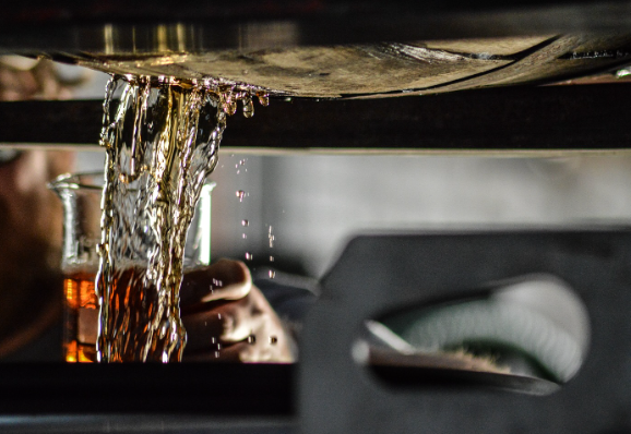 A close-up photograph capturing a rye whiskey pouring from a barrel into a beaker. 