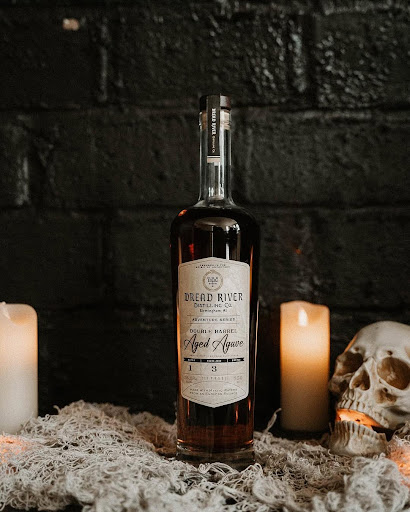 A bottle of Dread River Distilling Co.'s Double Barrel Aged Agave, set against a dark backdrop with candles and a skull, evoking a moody atmosphere.