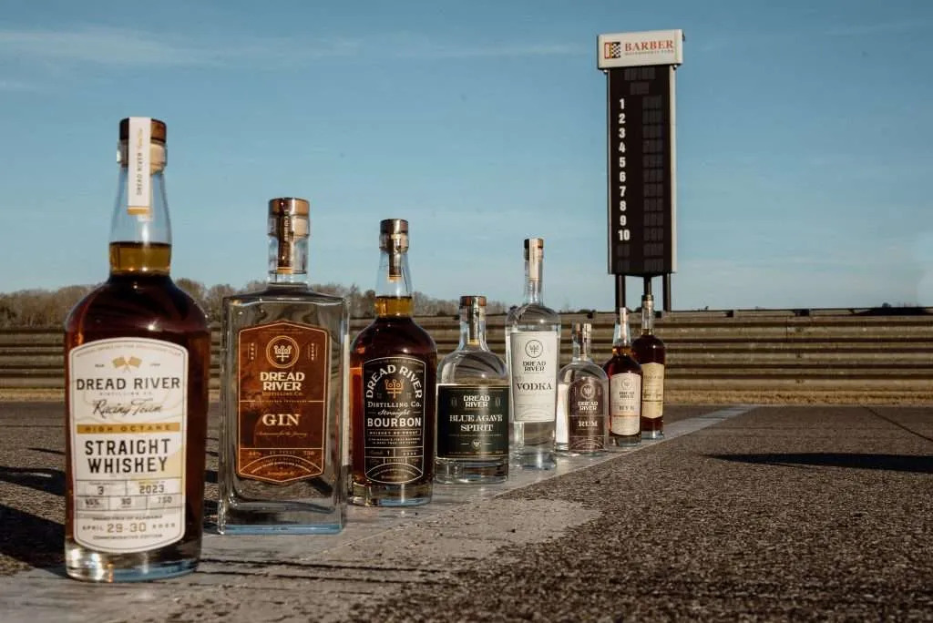 A lineup of Dread River Distilling Co. spirits, including whiskey, gin, bourbon, vodka, and rum, with a race track in the background.