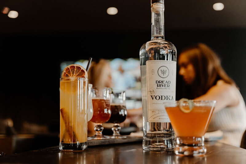 A bottle of Dread River Vodka displayed with various cocktails on a bar counter, with two people in the background.