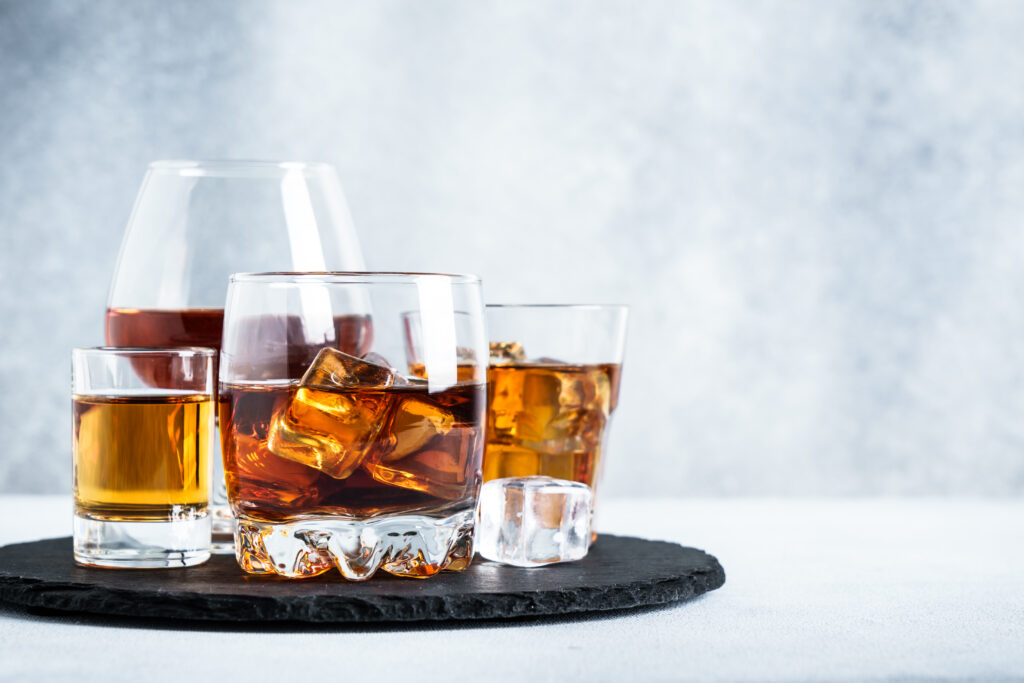 A variety of whiskey drinks in glasses with ice cubes on a slate coaster.