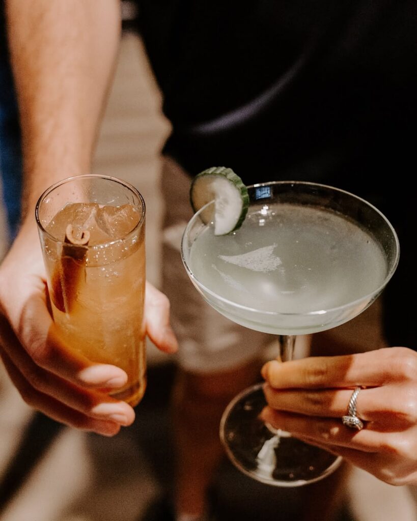 Two craft cocktails being served at a bar, including one with a cucumber garnish.