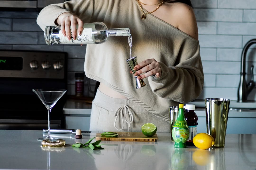 A woman pouring vodka into a jigger while preparing a cocktail at a home bar.
