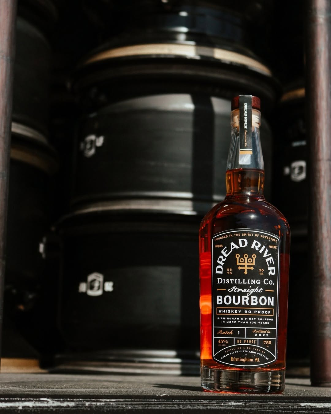 Dread River Distilling Co. Straight Bourbon bottle displayed in front of stacked whiskey barrels inside a rustic Birmingham distillery.