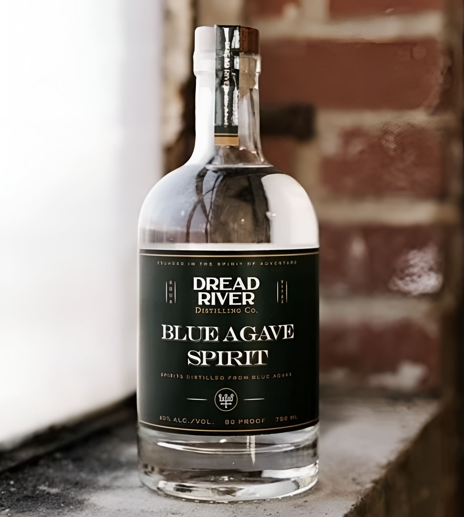Dread River Blue Agave Spirit bottle on a windowsill; green label in focus, brick wall, and daylight in background.