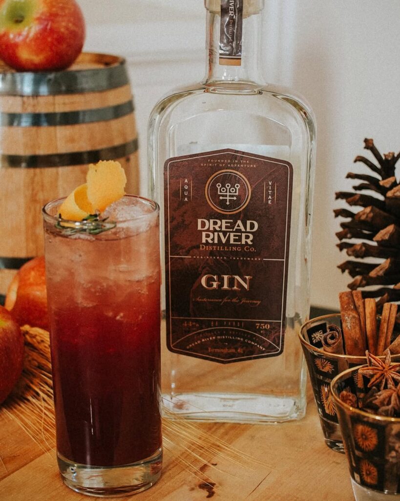 Dread River Gin bottle beside a tall iced gin cocktail with citrus twist, apples, and spices.