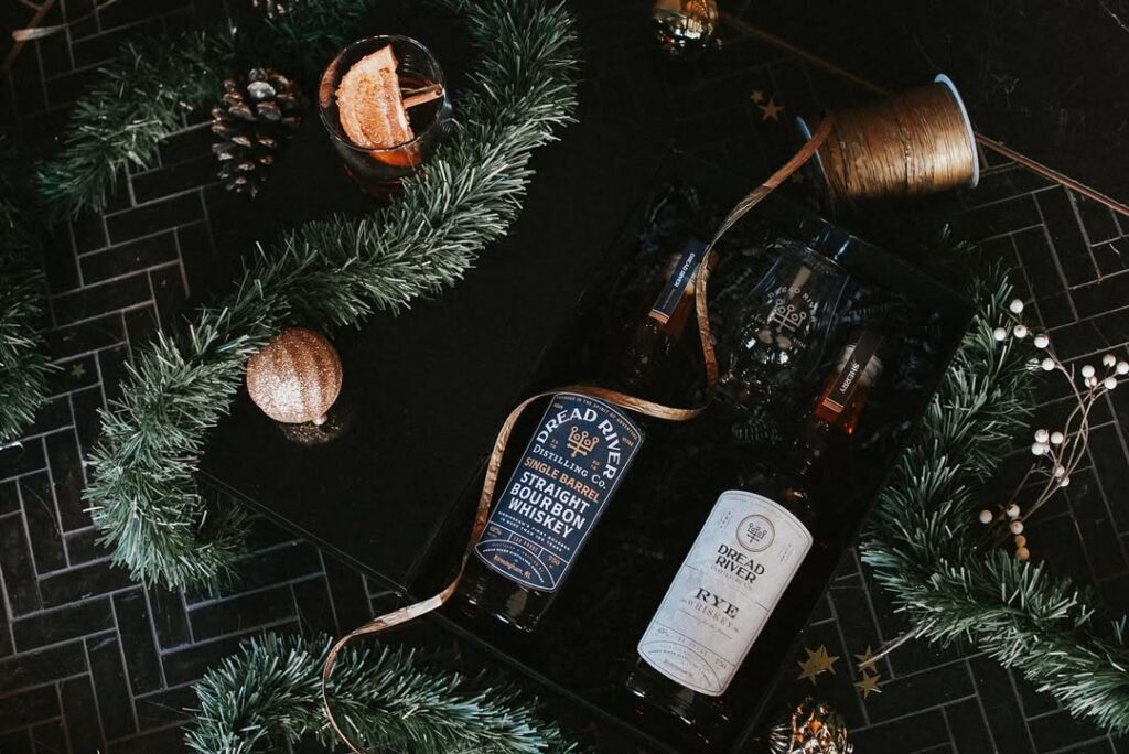 A bottle of Dread River Double Barrel Bourbon Whiskey and Dread River Rye Whiskey in a box, surrounded with christmas garlands, a golden bauble, a golden ribbon and pinecones.