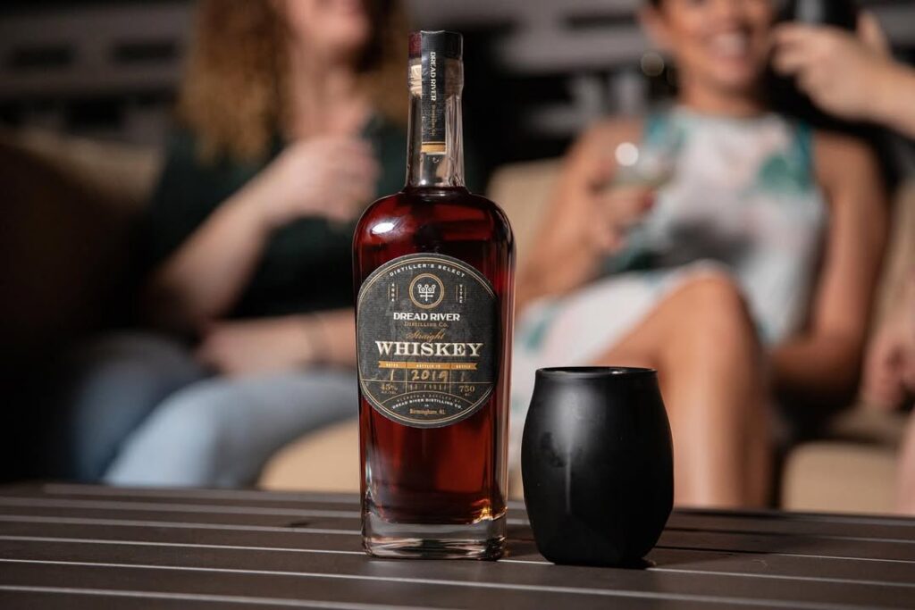 Dread River whiskey bottle on a table with guests enjoying drinks in a softly lit tasting room behind it.