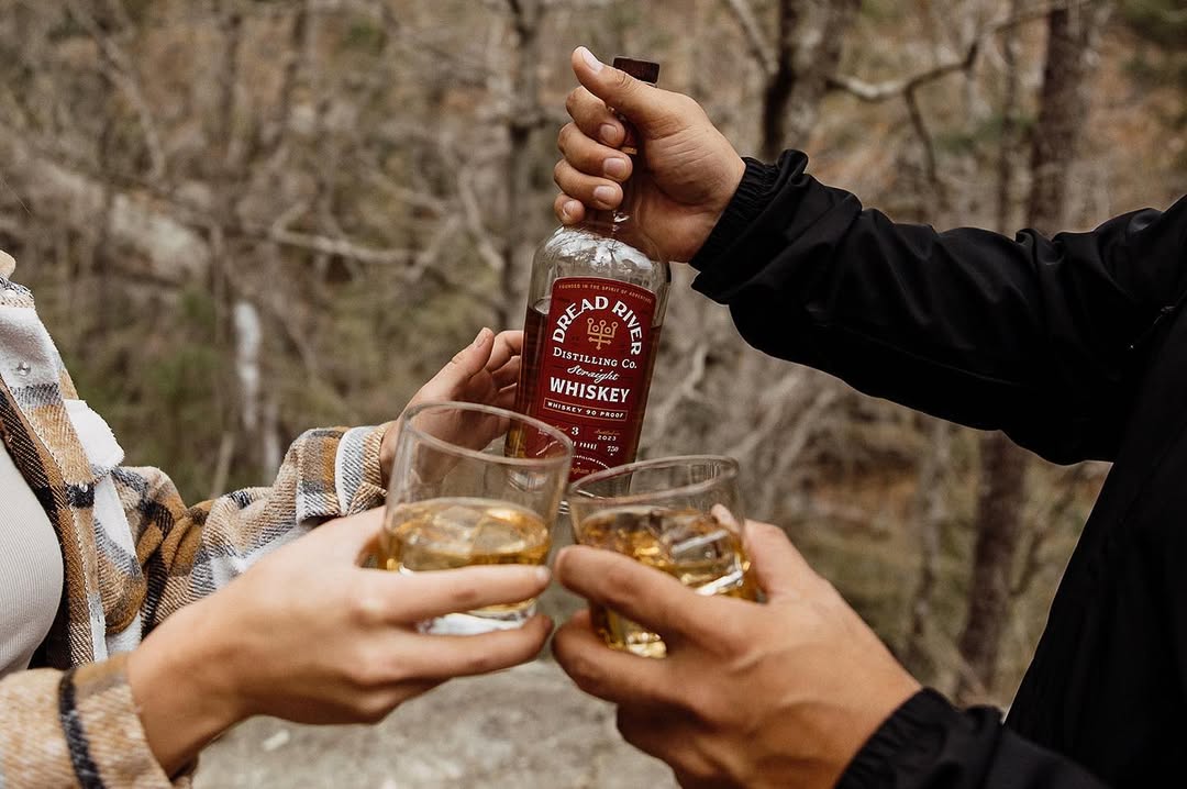 Friends toasting with Dread River whiskey glasses during an outdoor gathering surrounded by nature.