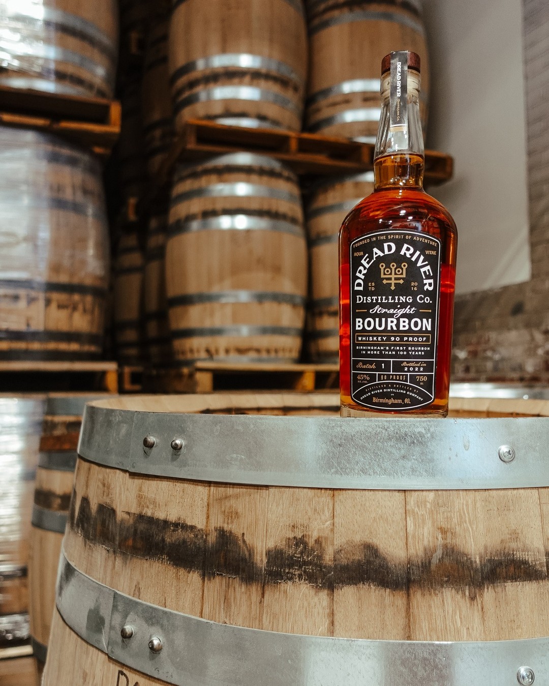 Bottle of Dread River Straight Bourbon resting on a wooden barrel with stacked aging barrels in the background.