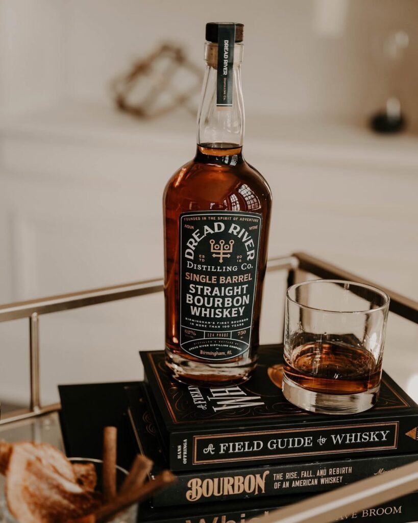 Dread River Single Barrel Bourbon bottle beside a whiskey glass and book on a wooden surface.