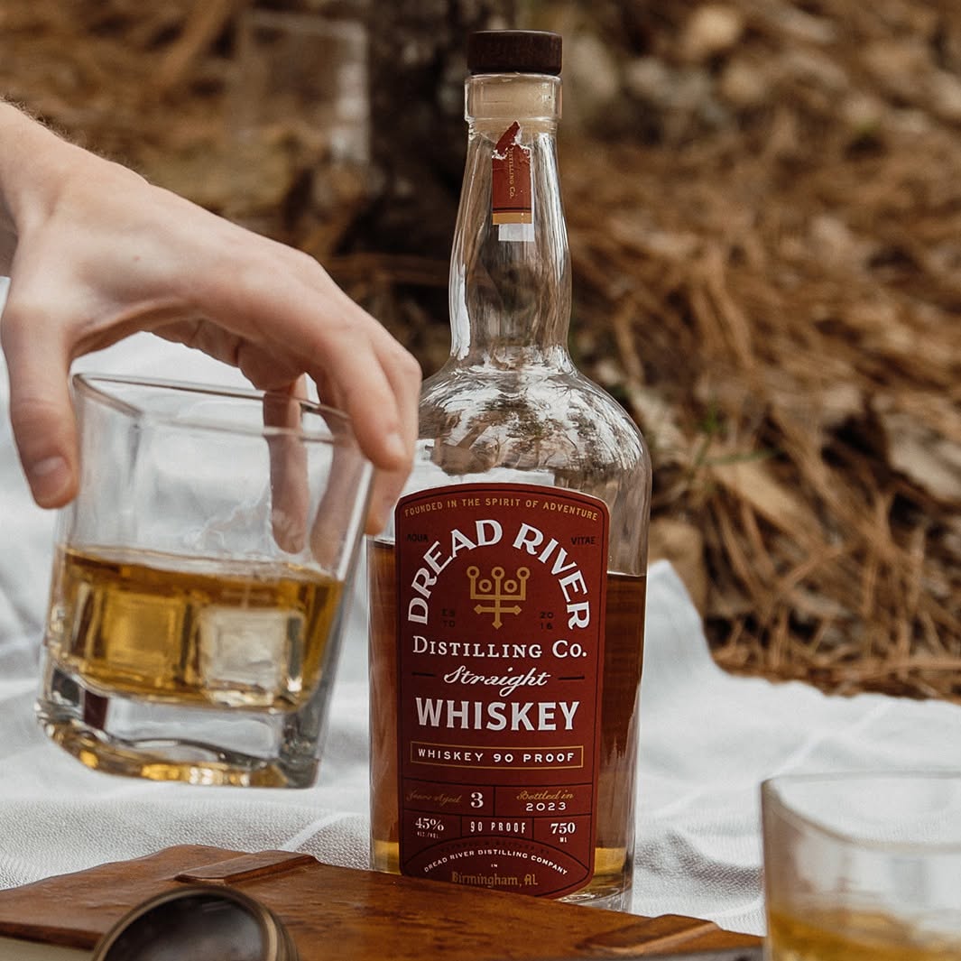 Bottle of Dread River Straight Bourbon Whiskey displayed outdoors beside two tasting glasses filled with whiskey.