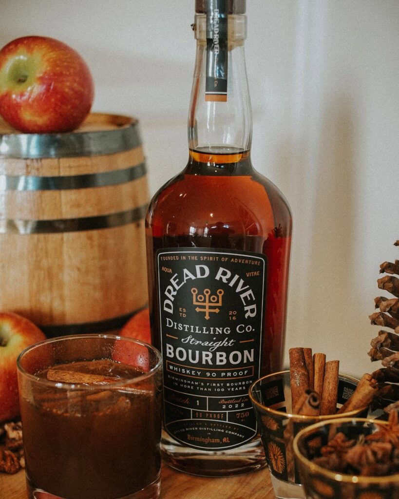 Dread River Straight Bourbon Whiskey beside an iced drink with apples, cinnamon sticks, and warm spices.