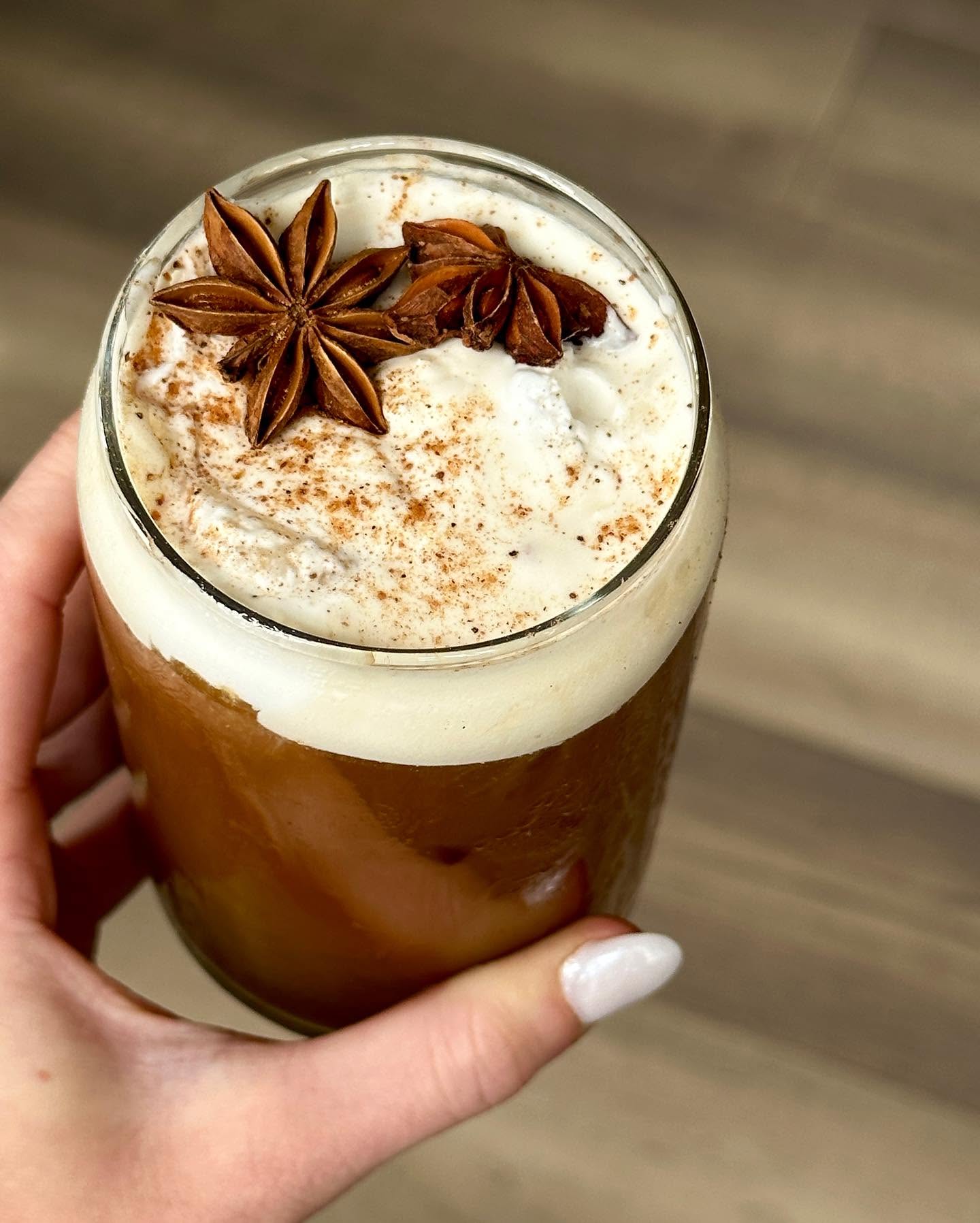 Hand holding a frothy espresso martini garnished with star anise and warm spices inside a modern cocktail bar.