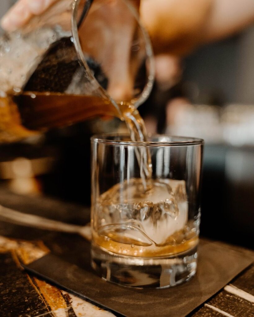 Dread River whiskey poured over a large ice cube in a rocks glass beside a glass pitcher.