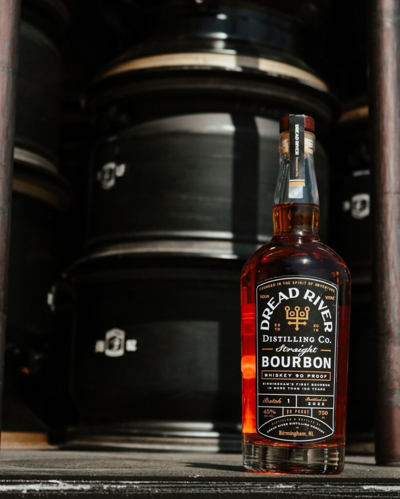 Dread River bourbon bottle positioned in front of large aging barrels in a distillery setting.